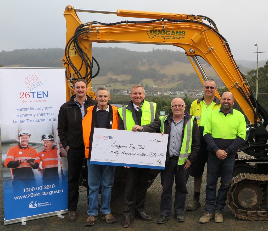 Workplace Grant project - Duggans. 
Photo of multiple people wearing high visibility vests, standing in front of a digger, and holding a giant 26Ten cheque