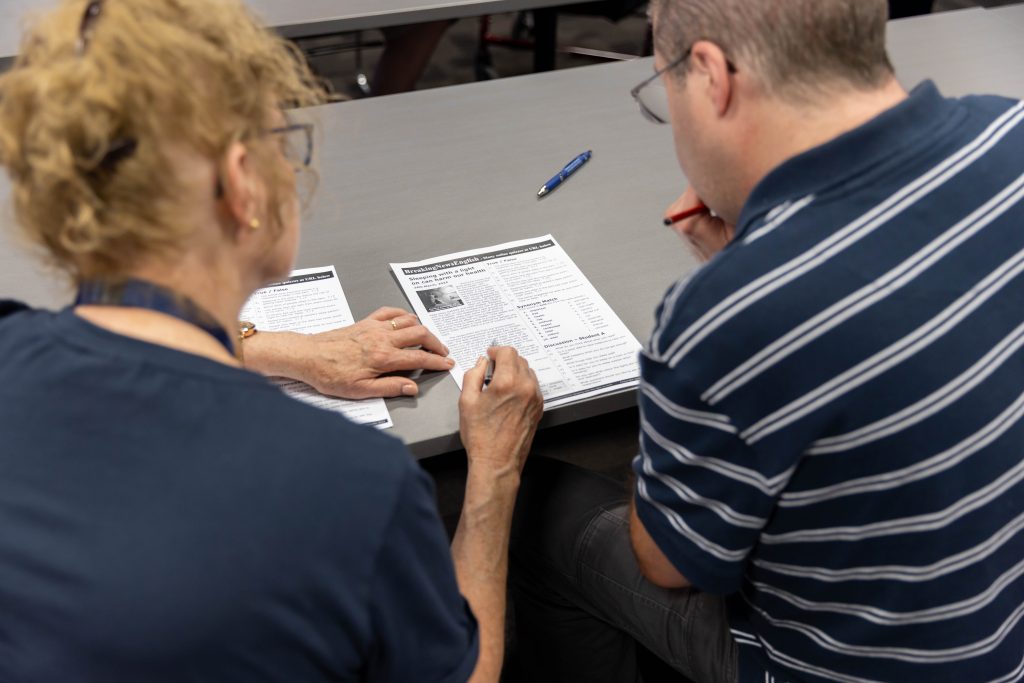 Photo of two people sitting at a table looking at an article on a piece of paper. Photo taken from behind