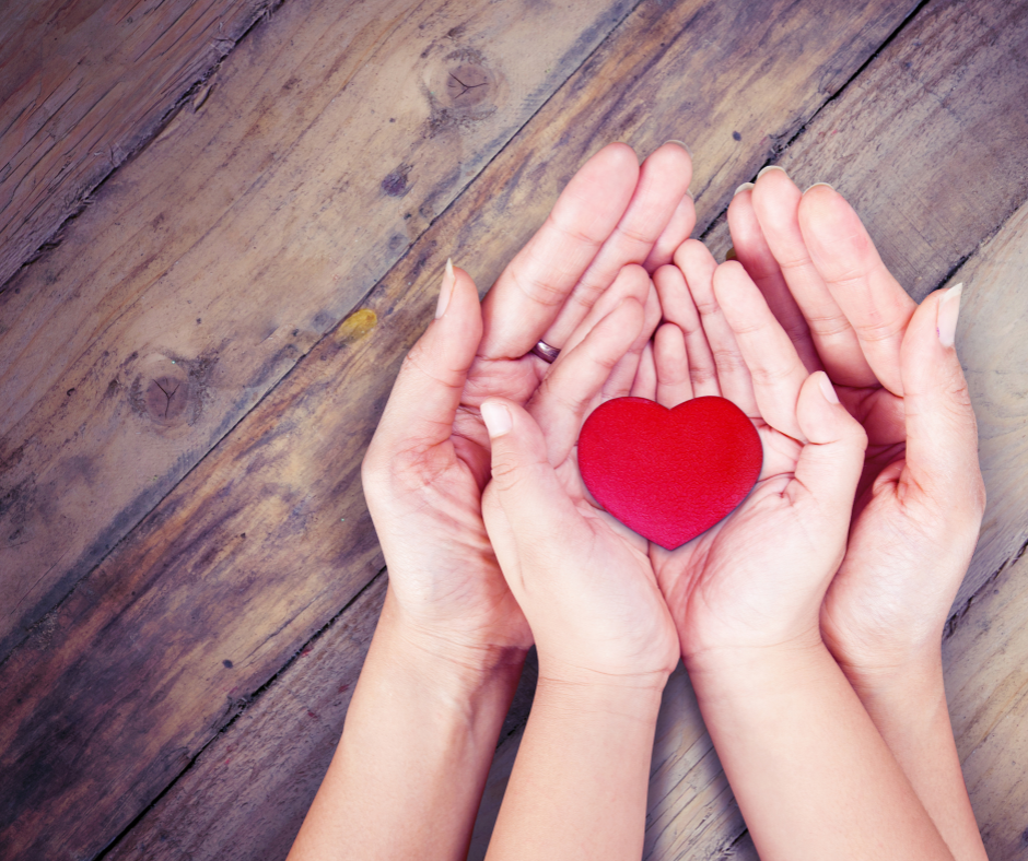 Photo of a child's hands holding a red love heart. The child's hands are cradled in an adult's hands