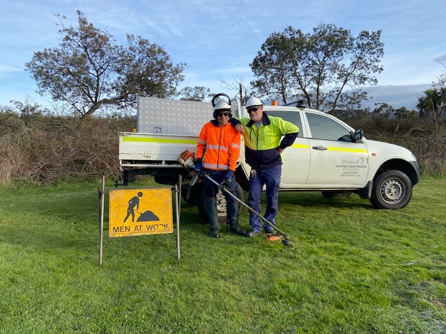 Photo of Bluegum Grounds Maintenance employees, Glenn and Alex, standing in front of a utility vehicle with a sign that says, 'men at work'. Both men are dressed in high visibility clothing.